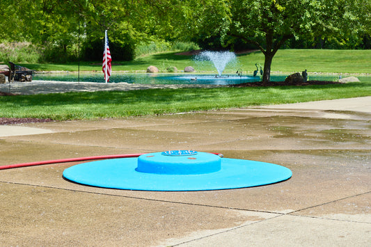My Splash Pad 5-foot portable splash pad in bright blue with raised center and spray nozzles, shown on a backyard patio with fountain and trees in the background.