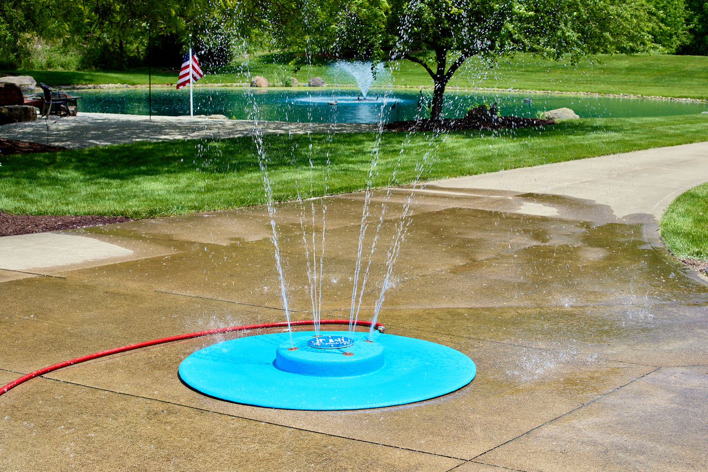 My Splash Pad 5-foot portable splash pad spraying water streams on a backyard patio with pond and fountain in the background.