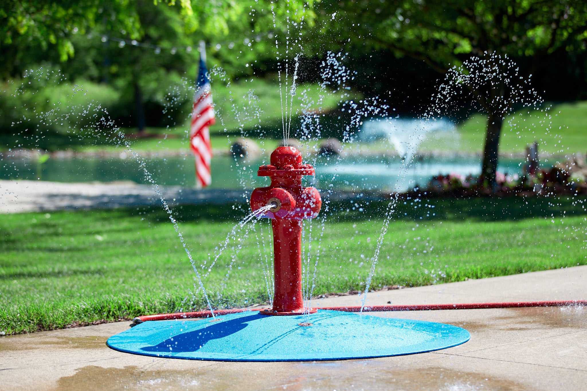 Red fire hydrant splash pad spraying water outdoors on a sunny day, with an American flag and pond in the background.