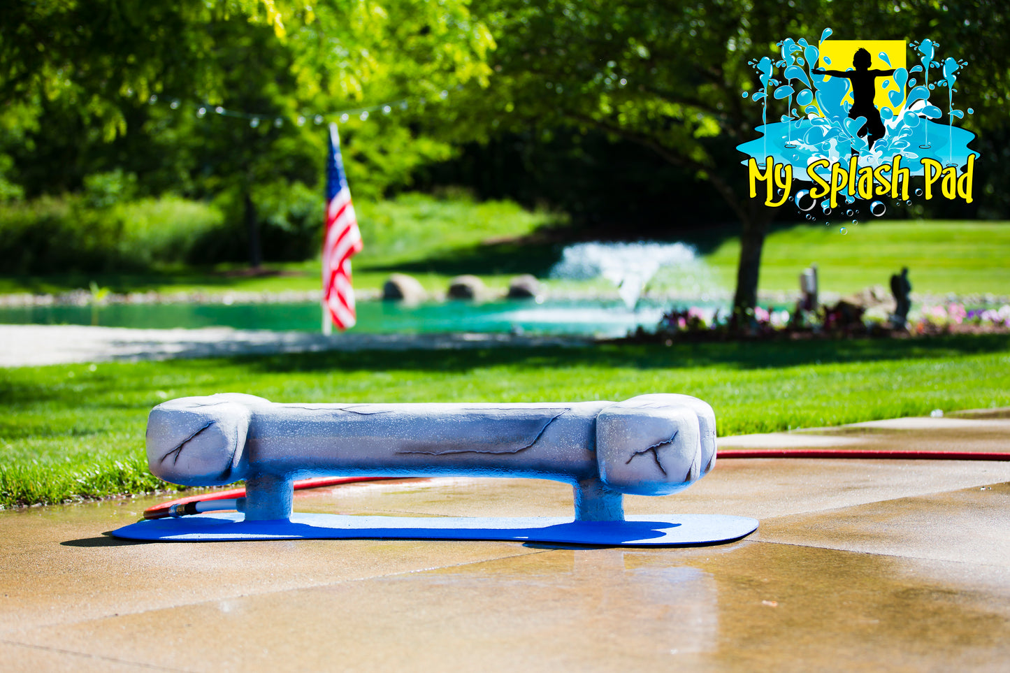 Dog Bone Water Play Feature by My Portable Splash Pad – sculpted gray dog bone splash toy on a blue base, outdoor setup on concrete with green lawn and pond in the background.