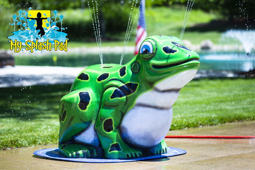 Green frog spray feature for My Splash Pad portable water play system, spraying water streams outdoors with pond and American flag in background.
