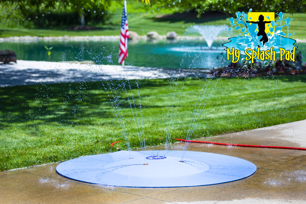 Round blue 7-foot portable splash pad spraying water upward on a sunny day, with American flag and pond in the background, My Splash Pad logo in the corner.