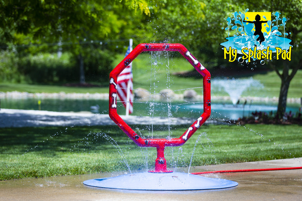 Red octagon dog jump hoop splash pad feature spraying water, part of My Splash Pad portable water play system, with pond and American flag in background.