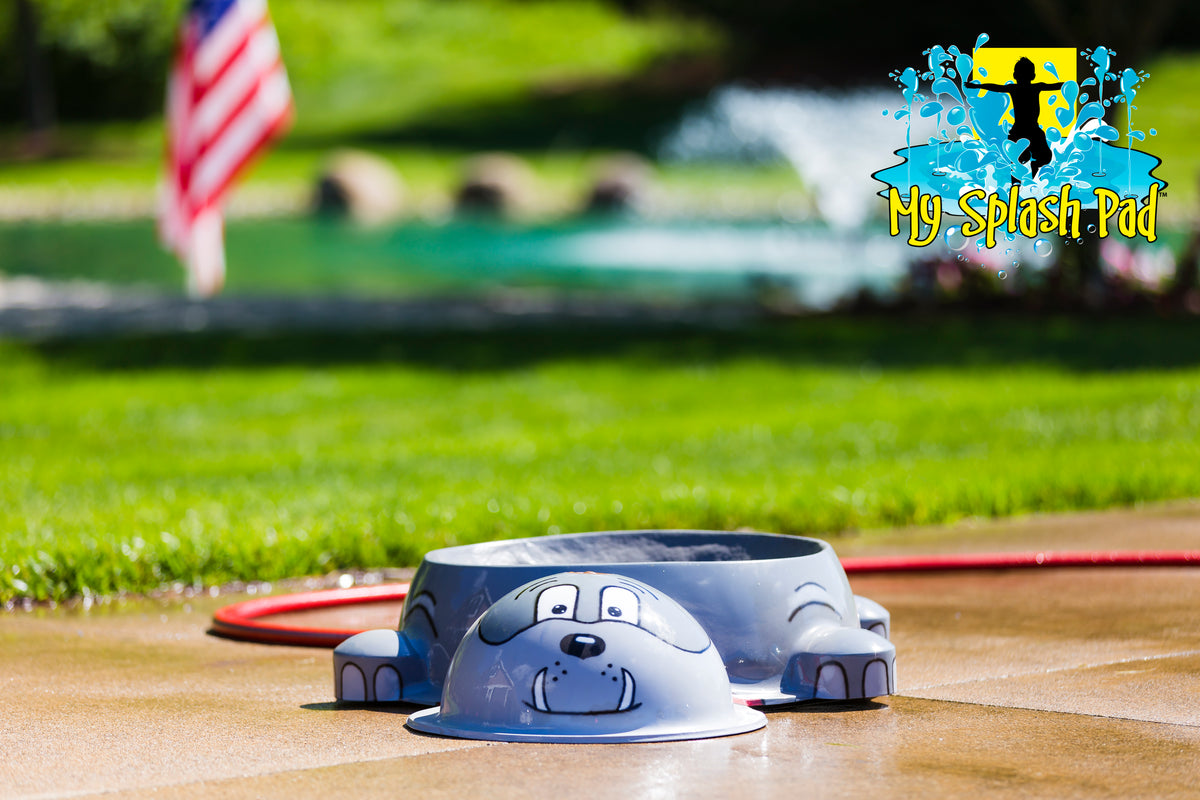 Dog Park Bulldog Water Bowl Water Play Feature by My Portable Splash Pad – gray bulldog-shaped water bowl on outdoor concrete with grass and American flag in background.