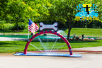 Doggy Wash Water Play Feature by My Portable Splash Pad – red arch with blue paw prints and white bone spraying water streams on outdoor concrete with grass, trees, and American flag in background.