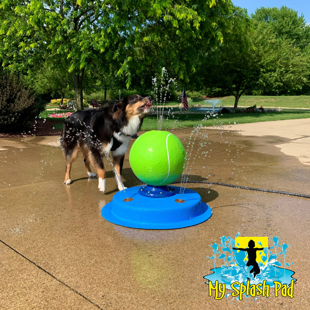 A portable splash pad featuring a green tennis ball at the top, with water spraying from multiple nozzles on a blue fiberglass base, placed on a concrete surface outdoors.